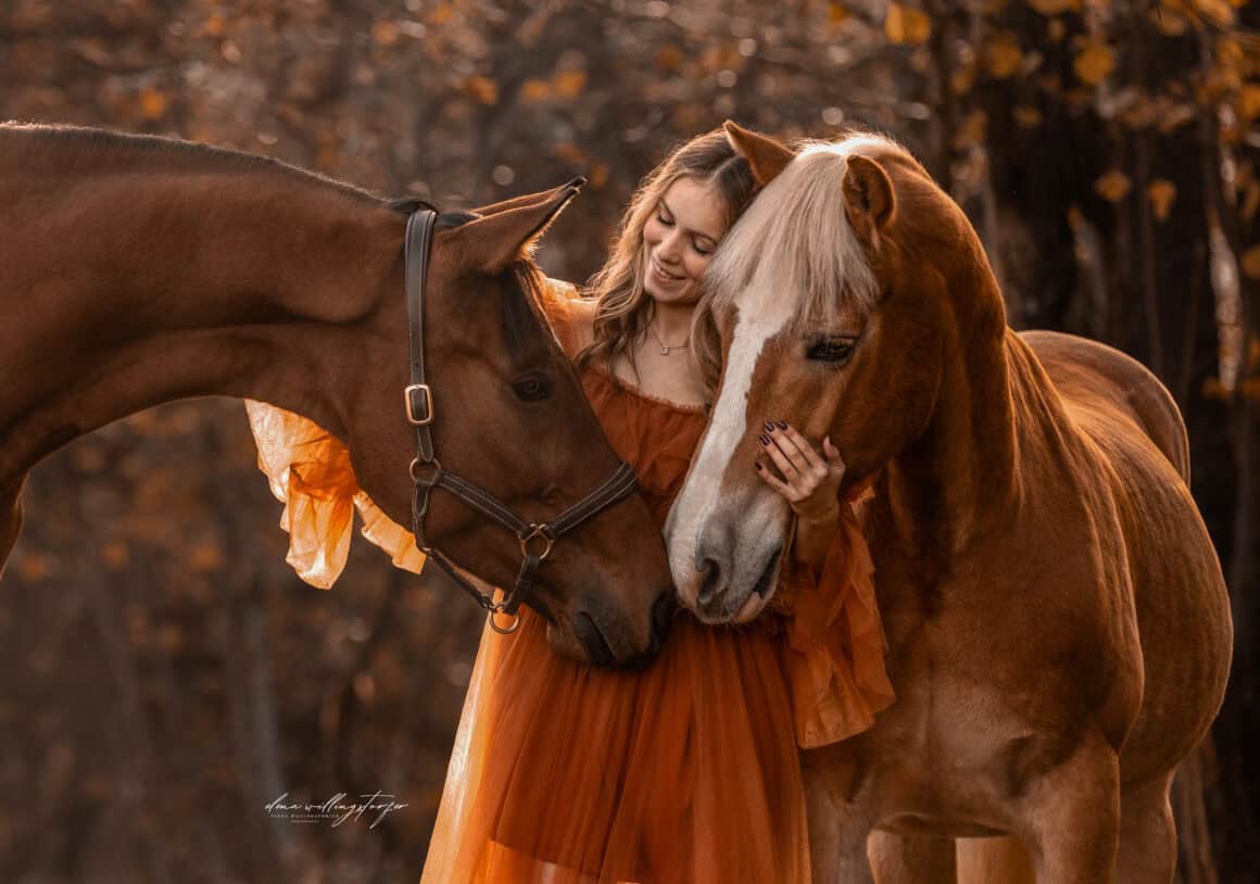 Carina Auer umarmt liebevoll ihre beiden Pferde Aquarell und Pop Up in einer herbstlichen Waldkulisse. ©️ Carina Auer / Elena Willingstorfer