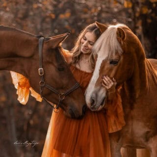 Carina Auer umarmt liebevoll ihre beiden Pferde Aquarell und Pop Up in einer herbstlichen Waldkulisse. ©️ Carina Auer / Elena Willingstorfer